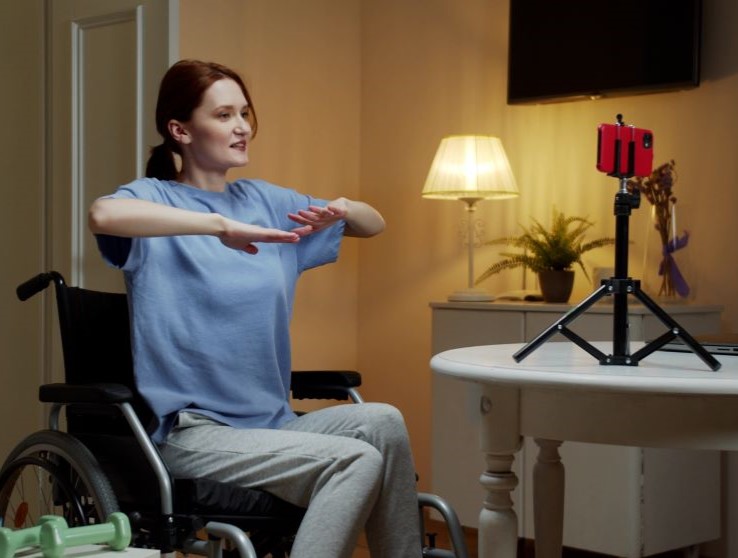 female wheel chair user watching phone on a tripod and holding up her arms to exercise